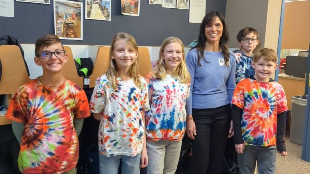 Sarah, a Global Doing Good Education Ambassador, stands with a group of students in Tie-Dye shirts in front of Roxbourgh Intermediate’s “Benjamin Wall”